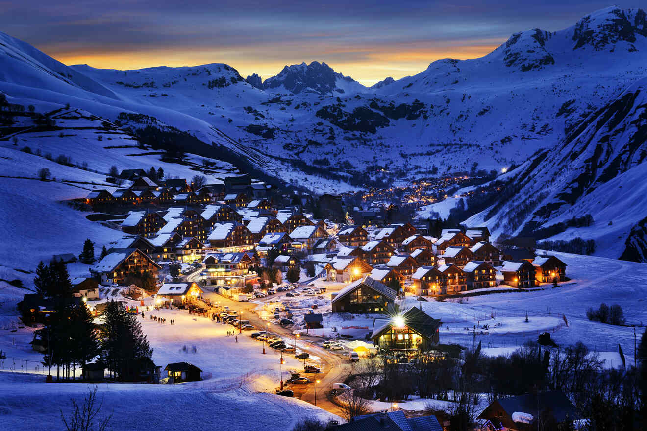 Snow-covered mountain village illuminated at dusk, with lights glowing from houses and streets, nestled among mountainous terrain under a twilight sky with a warm horizon glow.