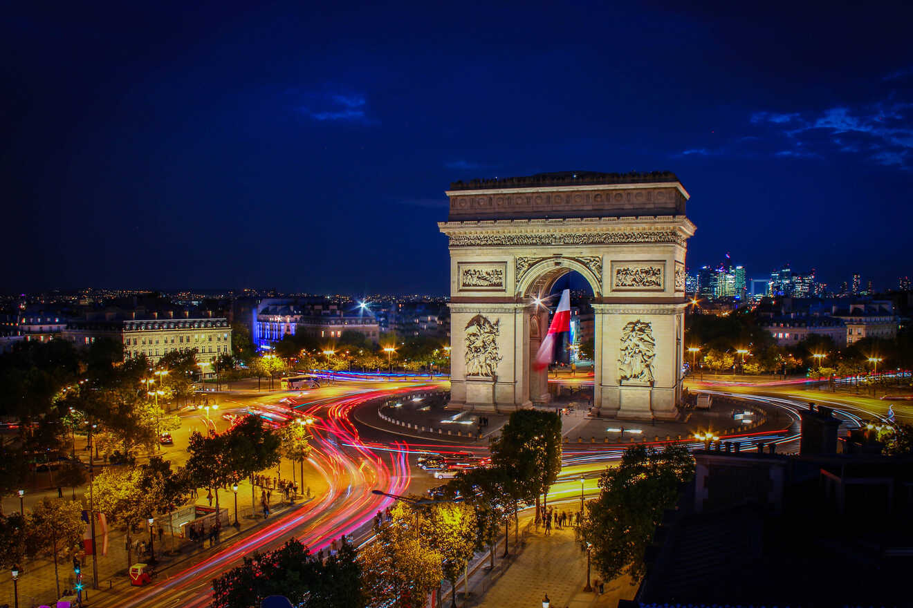 A nighttime view of the Arc de Triomphe in Paris, illuminated with traffic light trails circling around it, and the city skyline in the background.