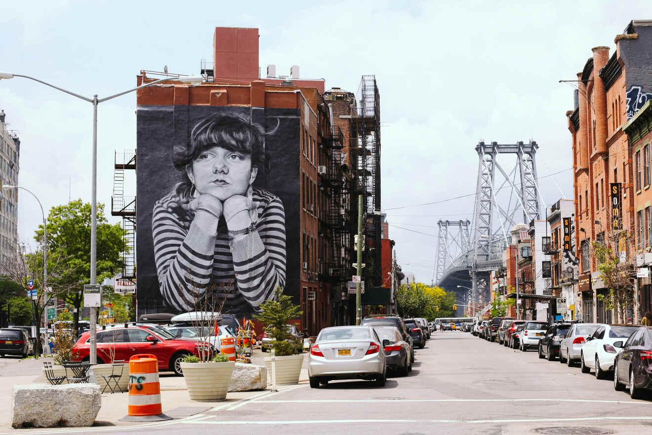 Street scene in a city featuring a large mural of a person on a building wall, with a bridge visible in the background and parked cars lining both sides of the road.
