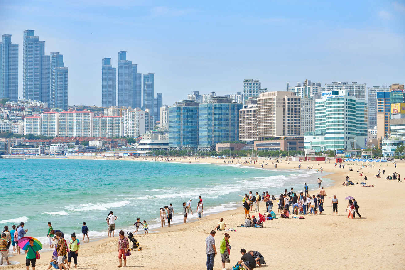 Busy beach scene with people enjoying the sandy shore and turquoise waters, skyscrapers in the background.