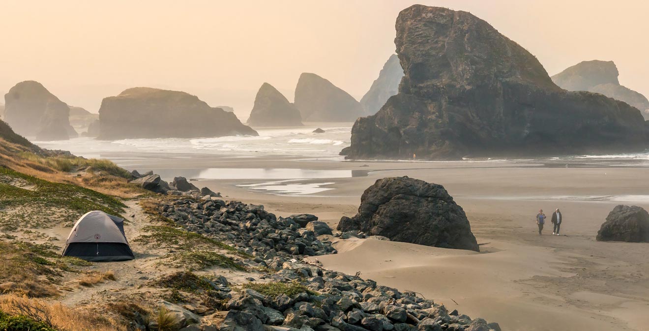 A tent is set up on a rocky beach with large sea stacks in the background. Two people are walking near the shoreline under an overcast sky.