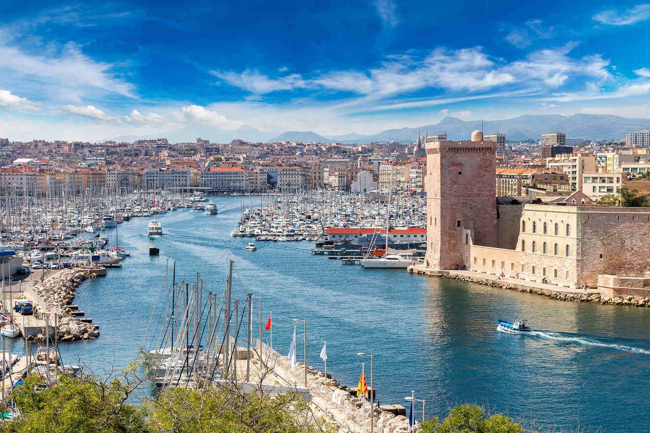 The historic Vieux-Port of Marseille with boats moored in the harbor, Fort Saint-Jean standing guard, and a backdrop of the city's terracotta rooftops and distant mountains