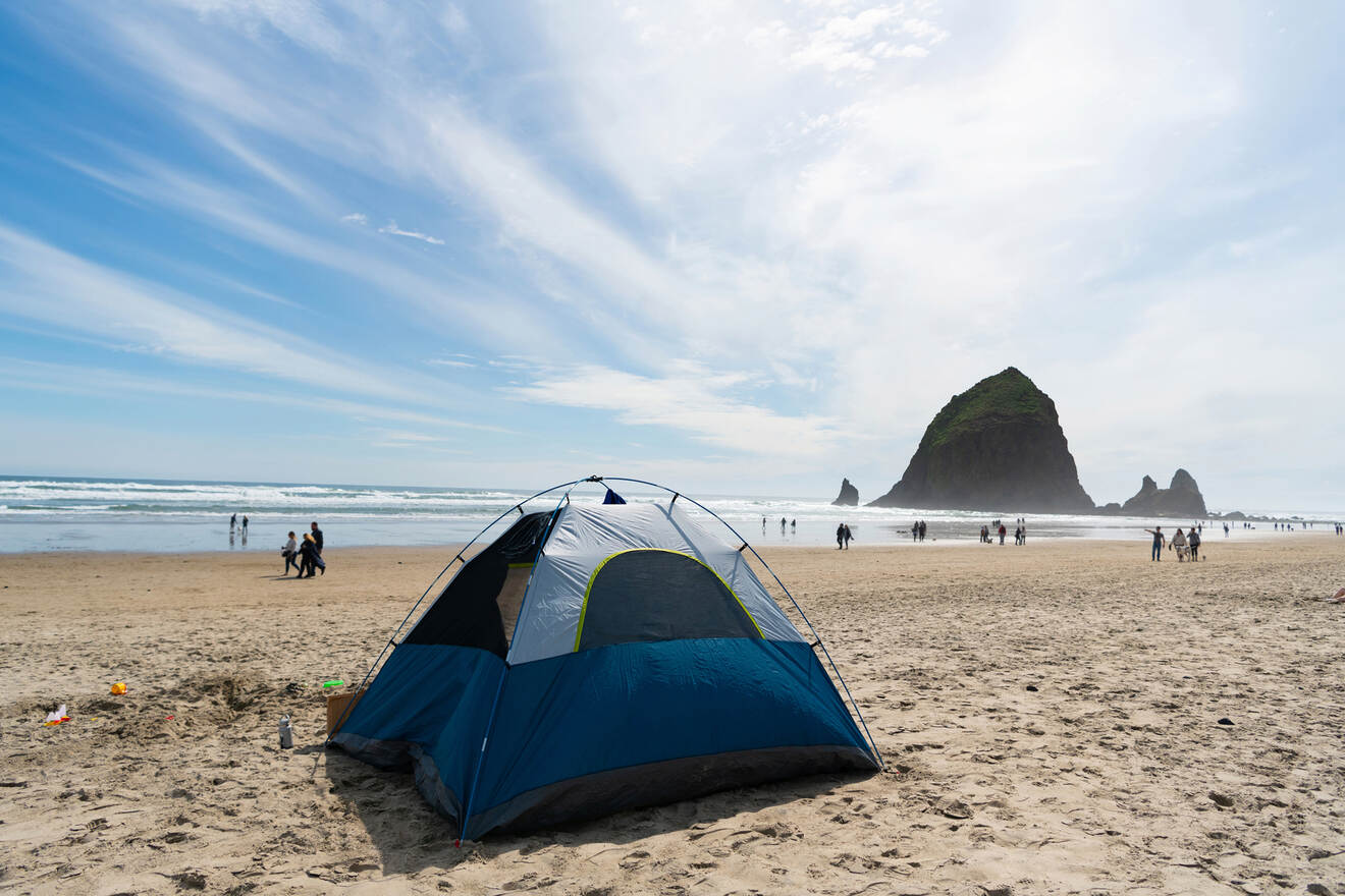 A blue camping tent set up on a sandy beach with people walking in the background and a large rock formation visible near the water's edge.