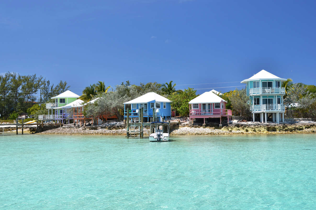 Colorful stilt houses line the shoreline against a backdrop of clear turquoise water and a vibrant blue sky. A small boat is moored by the shore. Trees and bushes surround the area.