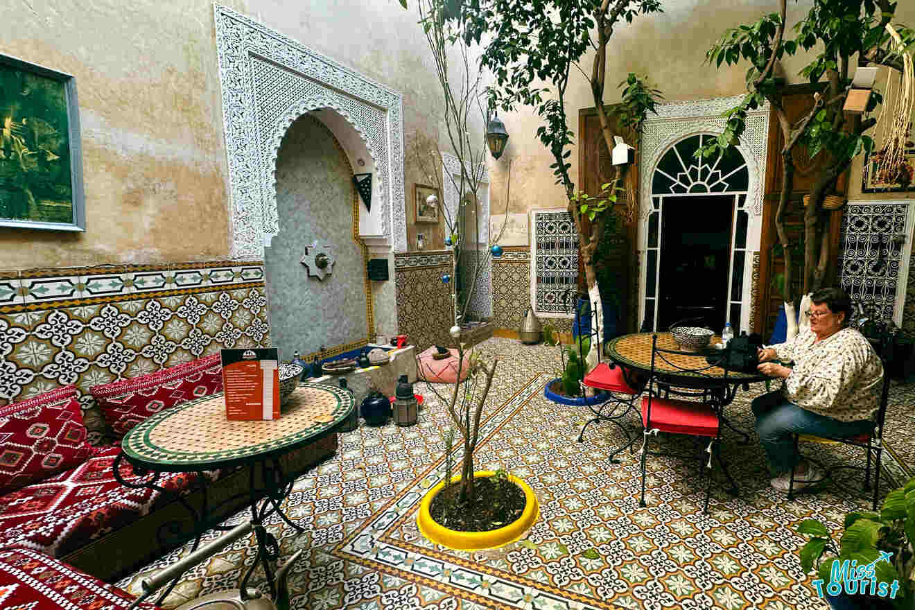 A woman sits in a mosaic-tiled Moroccan riad courtyard, surrounded by plants and traditional decor.