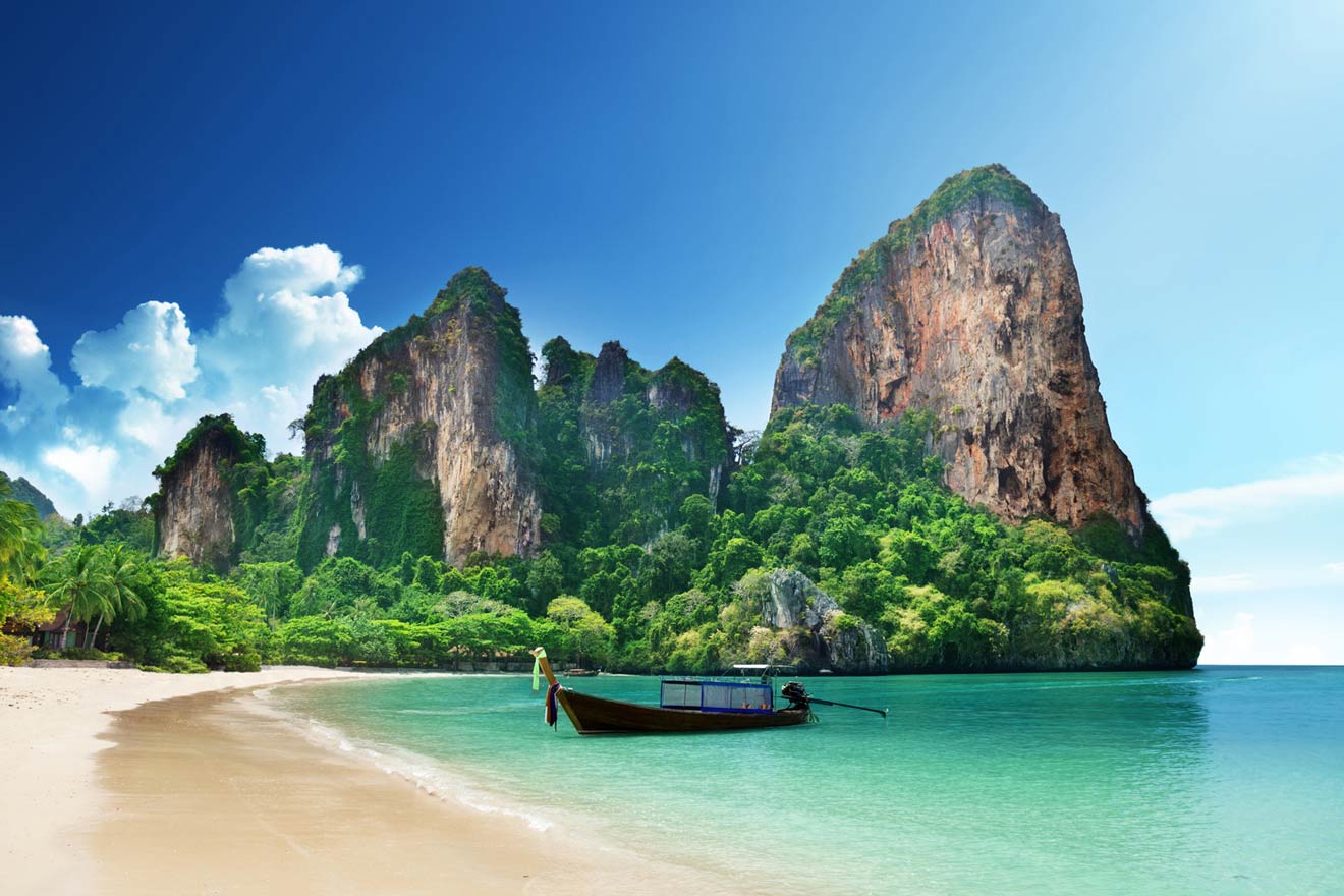 Scenic view of a pristine tropical beach with crystal-clear turquoise waters, a traditional longtail boat anchored on white sand, and imposing limestone cliffs under a bright blue sky