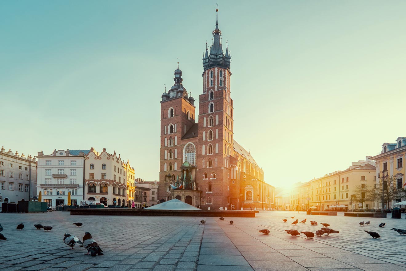 A historic church with two towers stands in a sunlit square with scattered pigeons and surrounding buildings.