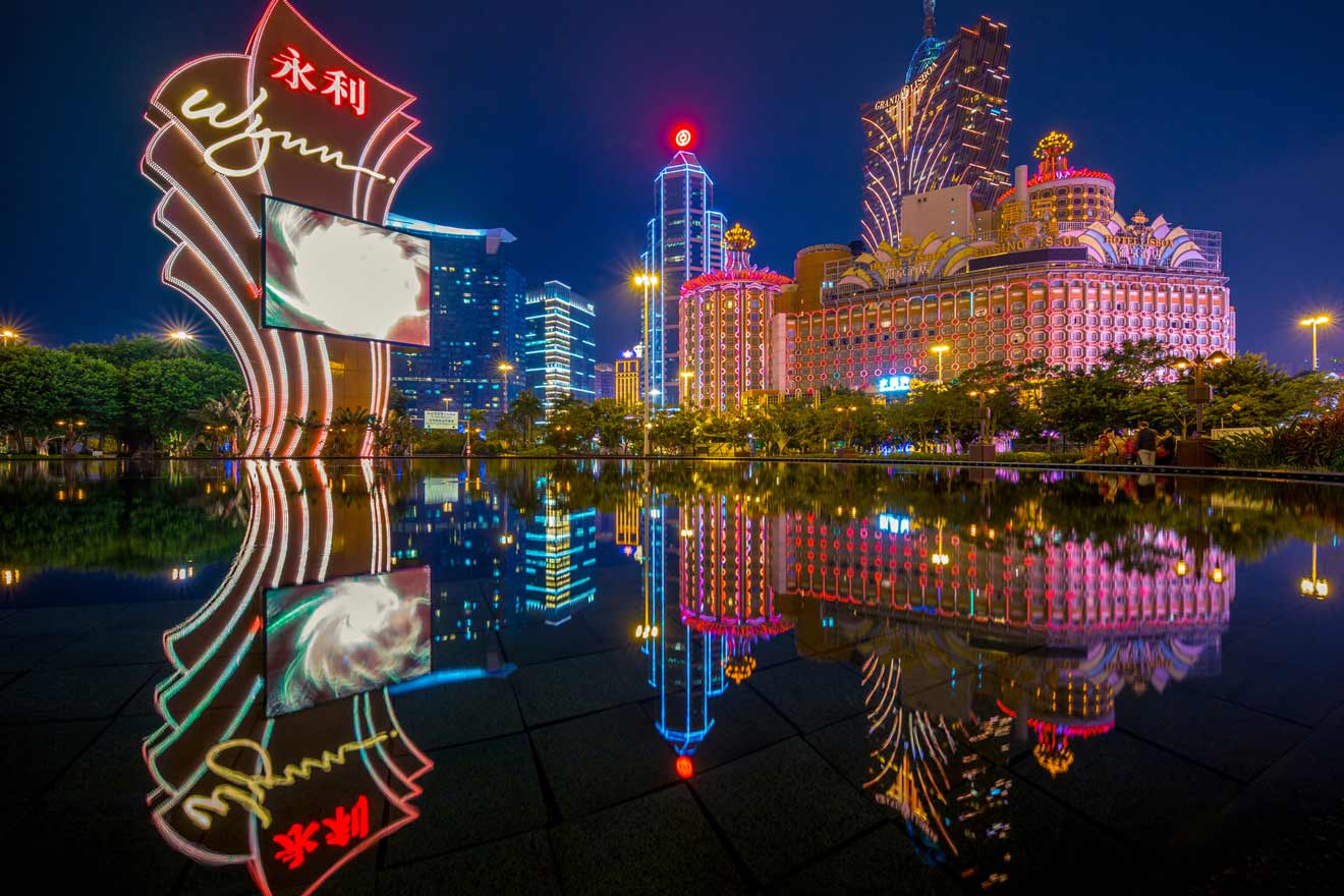 Night view of neon-lit casinos in Macau reflecting in a still pond, featuring vibrant signage from Wynn and Grand Lisboa.