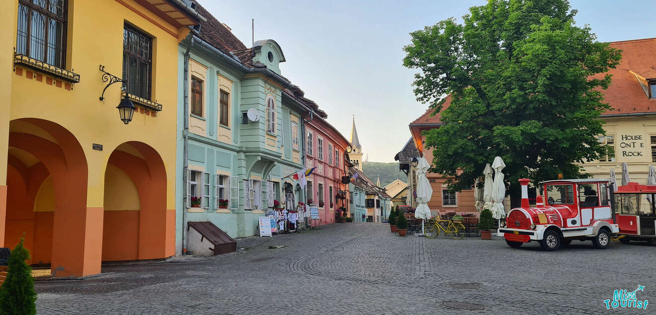 A quiet, cobblestone street with colorful buildings and a small red and white train. Table umbrellas are set up outside a building next to a large tree.