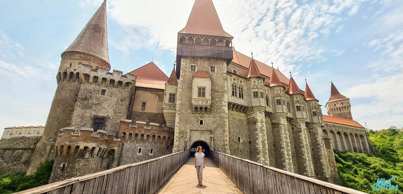 A person stands on a wooden bridge leading to a large medieval castle with multiple towers under a partly cloudy sky.