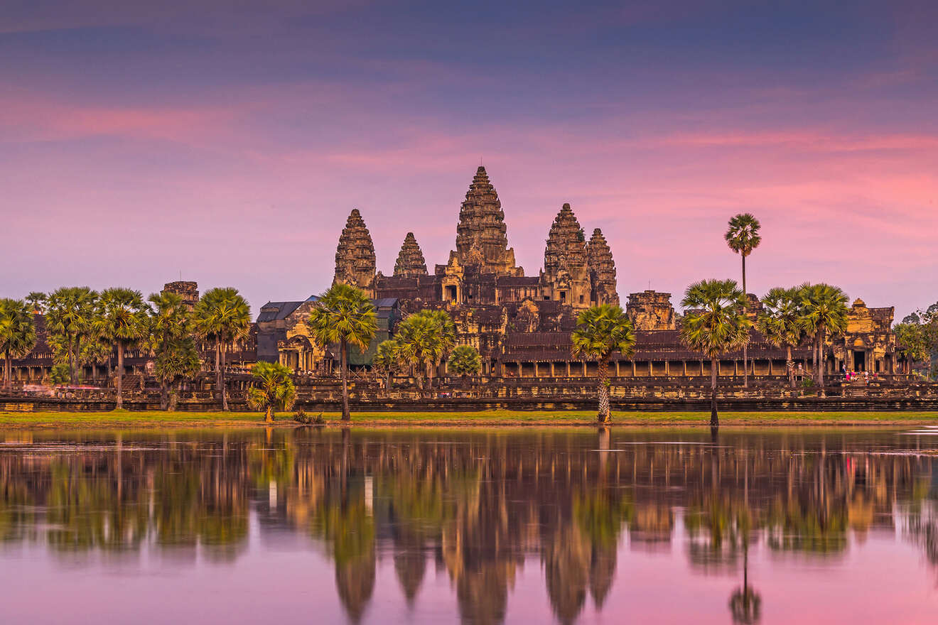 Sunset view of Angkor Wat temple in Cambodia, with its silhouette reflecting in the water, framed by tropical palms under a purple and orange sky