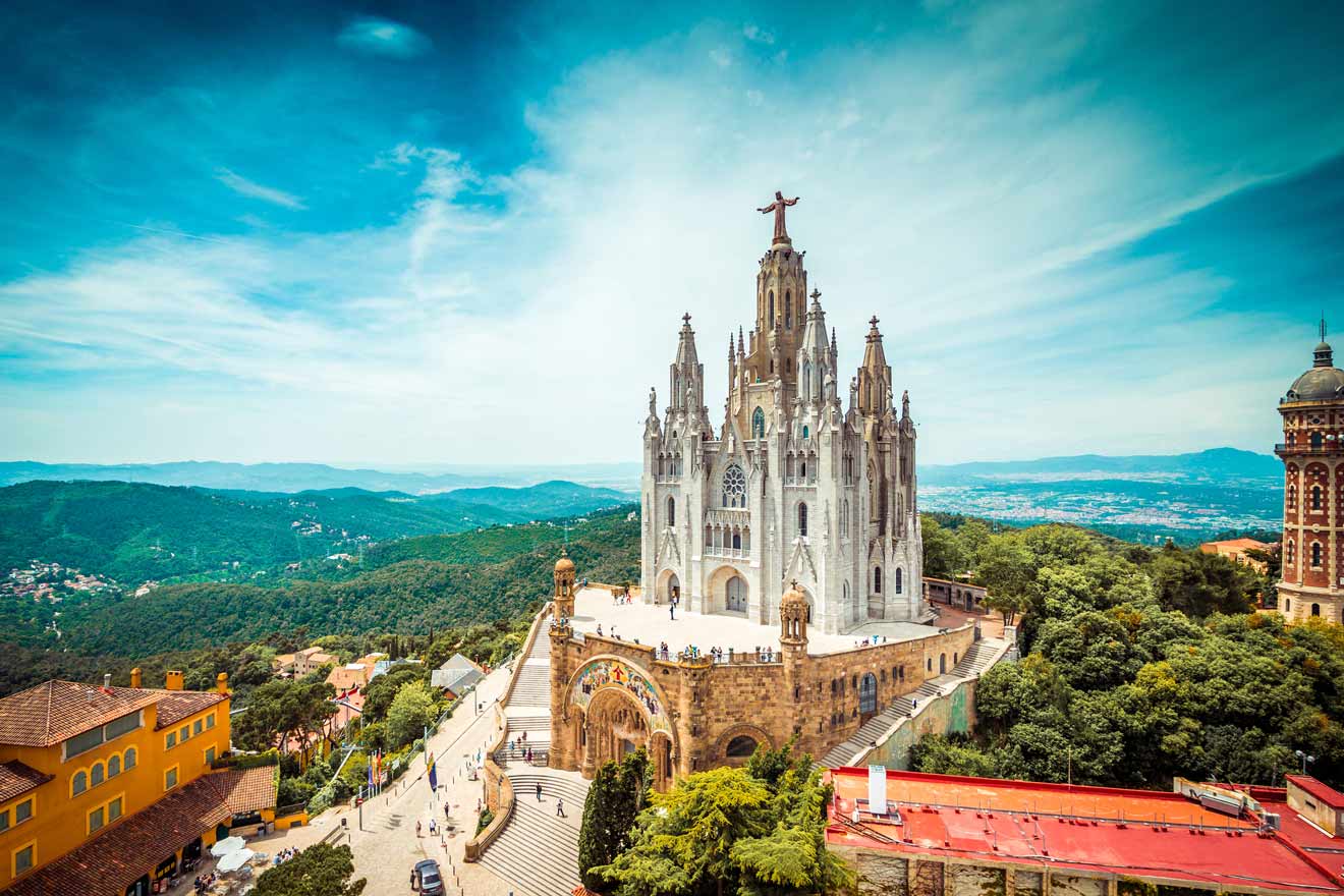 A large, ornate church with multiple spires sits atop a hill surrounded by lush greenery, with a distant mountainous landscape under a partly cloudy sky. Nearby, a red-roofed building and a tower are visible.