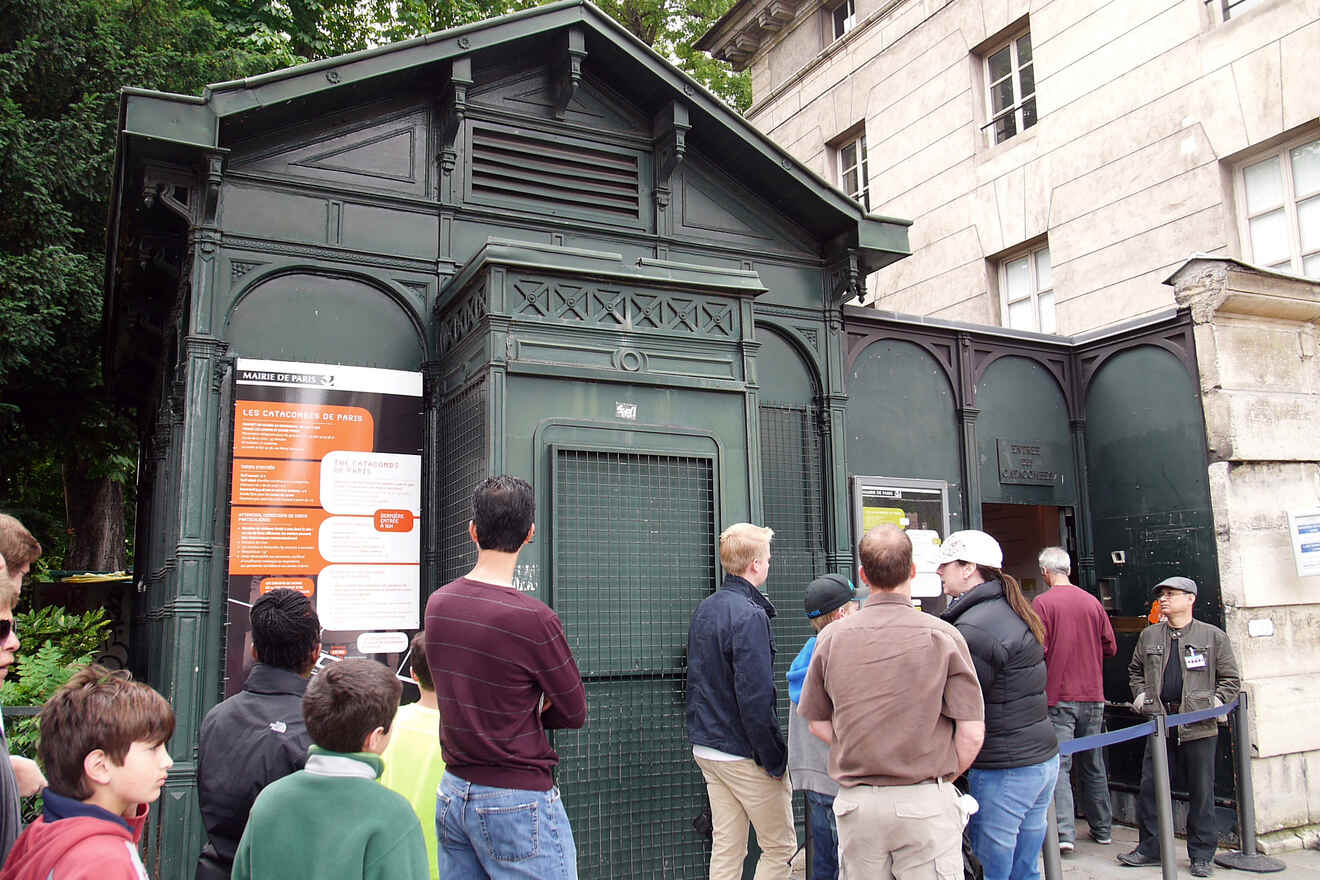 People stand in line outside a public restroom in an urban area, with informational signs on the kiosk's wall.