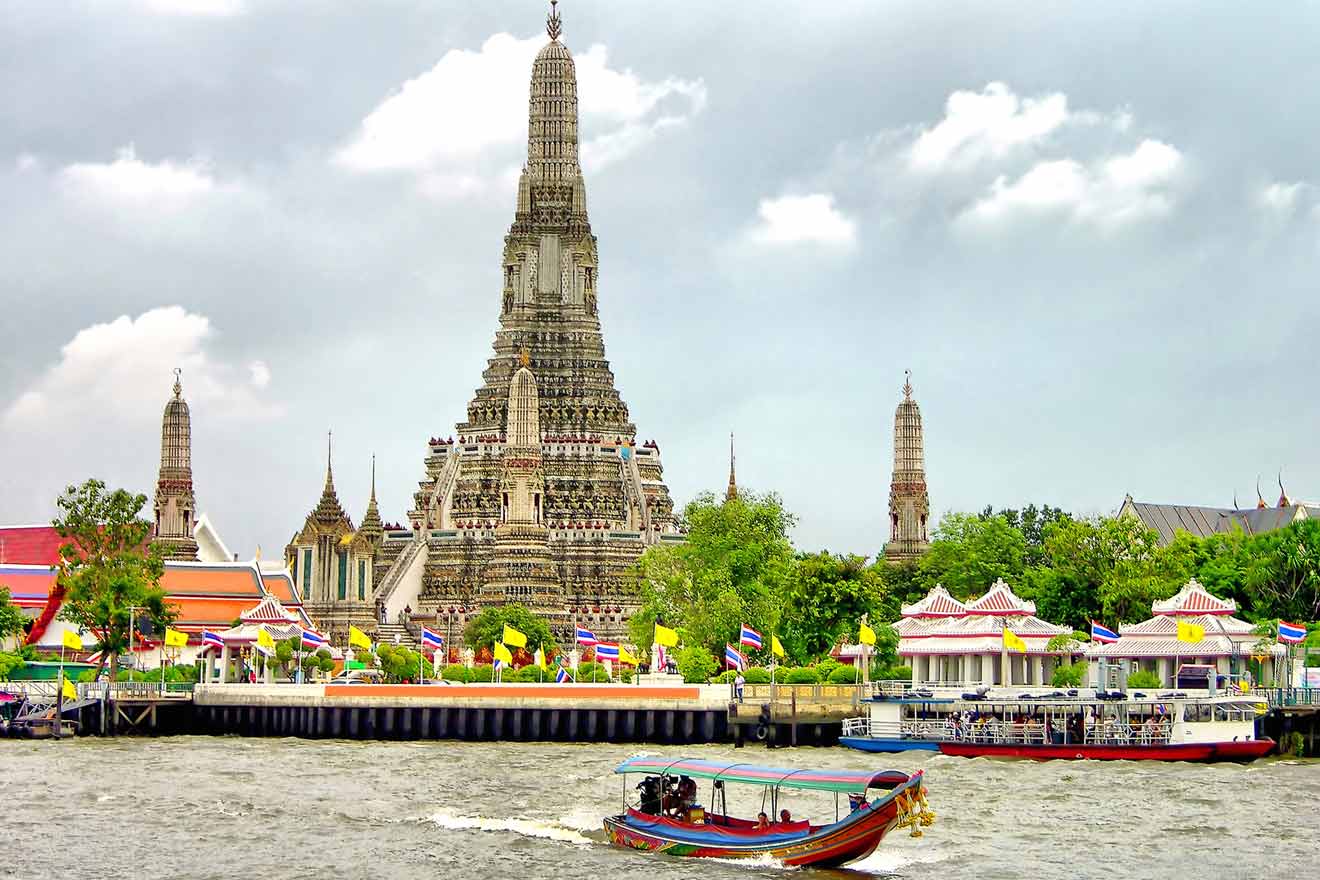 A colorful boat sails on the river in front of the ornate Wat Arun temple adorned with flags, set against a cloudy sky in Bangkok, Thailand.