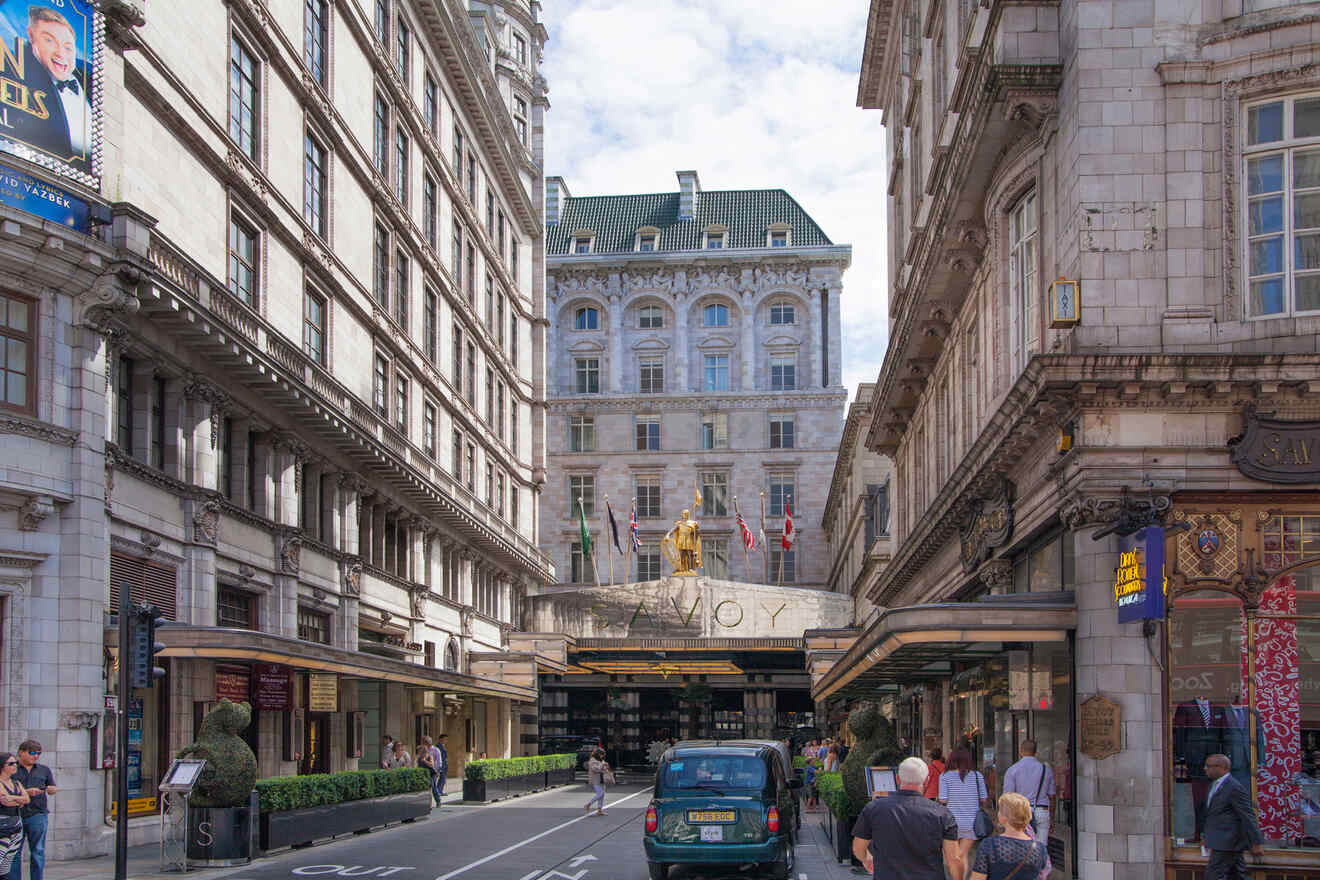 A view of the entrance to the Savoy Hotel in London, with a line of vehicles and pedestrians on the street, and a statue visible above the entrance.