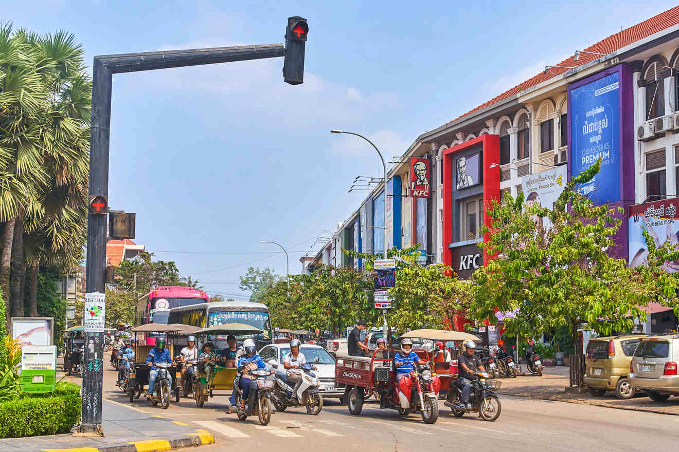 Bustling street scene in Cambodia with a red light at the crosswalk, motorbikes and tuk-tuks waiting, and a row of buildings with colorful signage including KFC