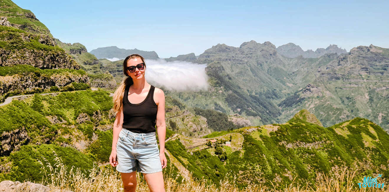 The writer of the post in sunglasses and casual clothing stands on a grassy hill with a scenic backdrop of mountains and clouds.