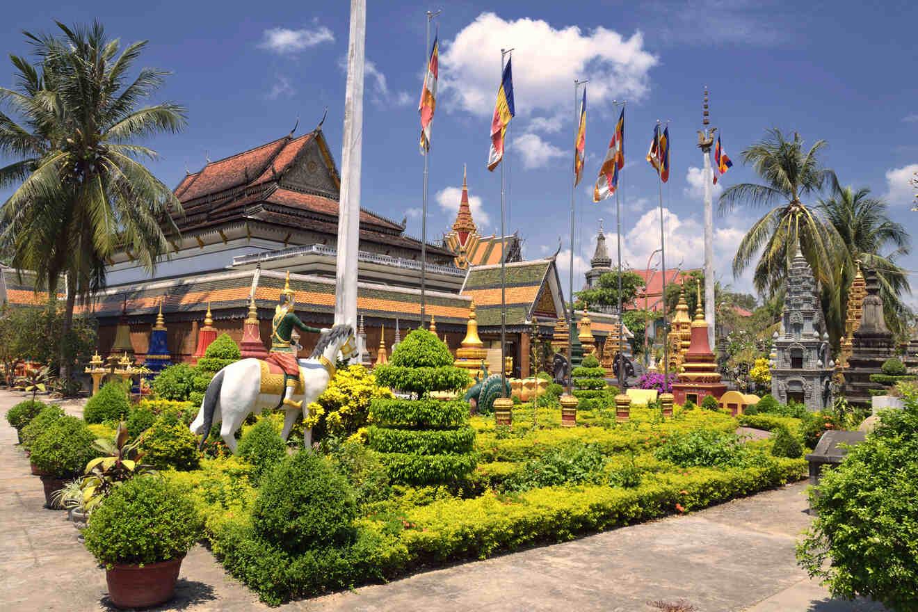 Lush garden in front of a Cambodian temple with ornate statues and vibrant national flags fluttering against a clear blue sky