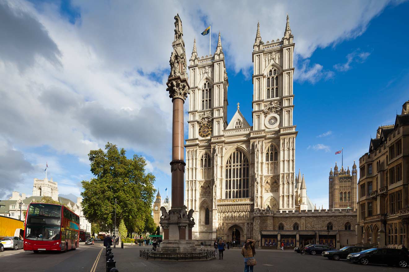 Westminster Abbey, the historic Gothic church in London, under a cloudy sky with the intricate facade visible and people and traffic in the foreground