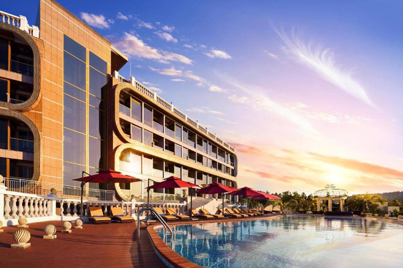 Modern hotel with a large outdoor swimming pool, sun loungers, and red umbrellas at sunset. The hotel features reflective windows and a distinct architectural design.