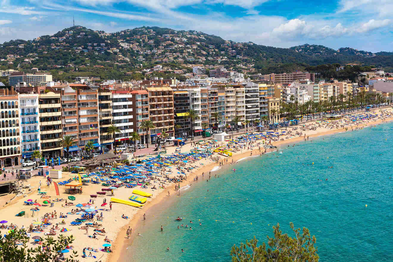 A crowded beach with colorful umbrellas and sunbathers, bordered by a row of multi-story buildings and a lush green hill in the background.