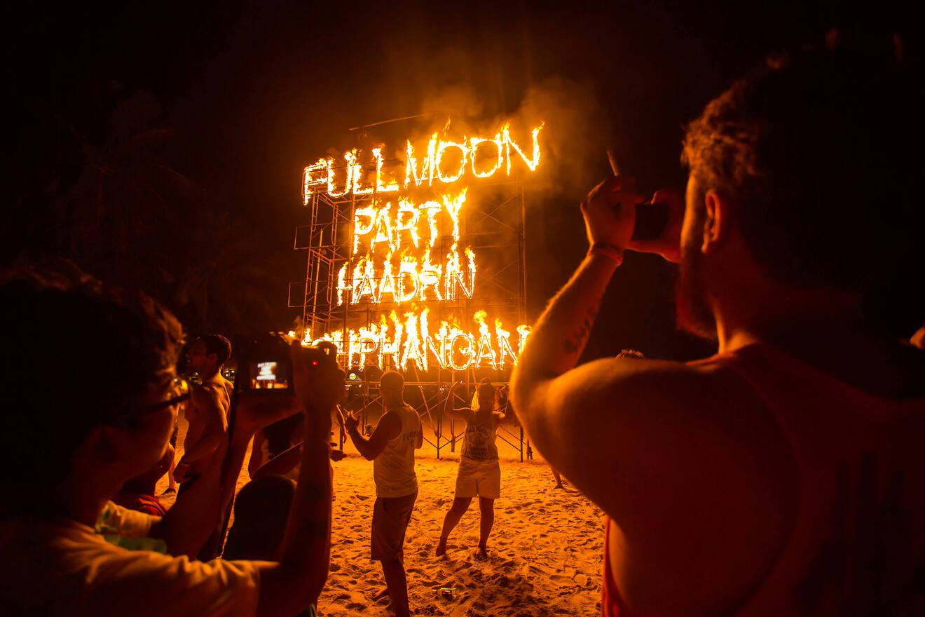 Crowd of people at night on a beach, captivated by a fiery display spelling out 'Full Moon Party Haad Rin Koh Phangan', with a silhouette of a person taking a photo in the foreground