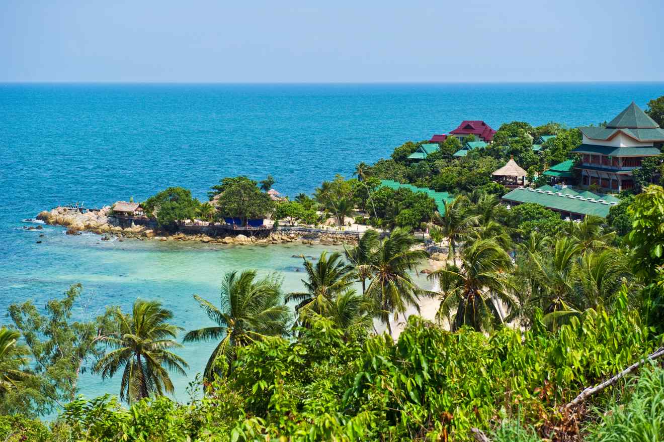 Scenic coastal view of a secluded cove on Koh Phangan, with traditional Thai architecture amidst lush tropical greenery, overlooking the clear blue sea