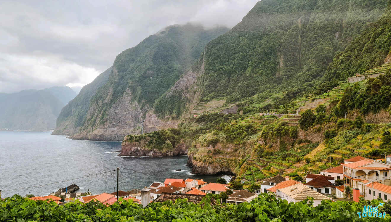 Coastal village with red-roofed houses nestled against steep, lush green cliffs, overlooking a rocky shoreline with misty clouds above.