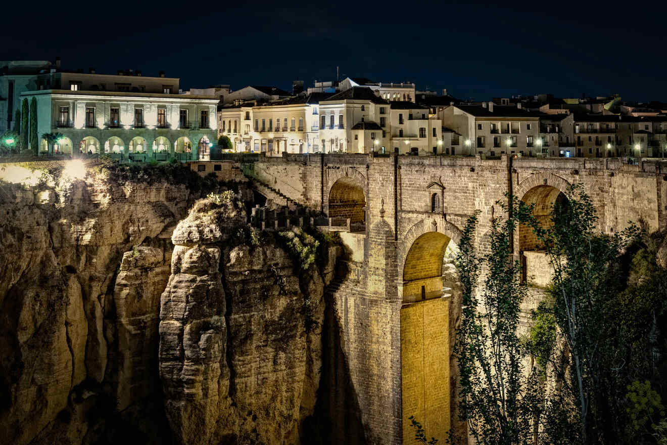 Night view of a historic stone bridge and adjacent buildings atop cliffs illuminated by lights.