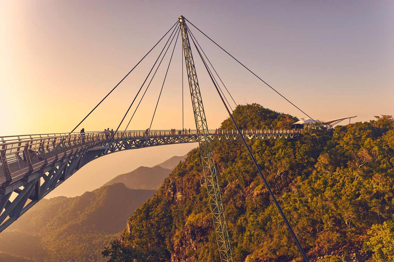 A large suspension bridge in Langkawi extending across a valley, with a mountainous landscape and a warm sunset sky.