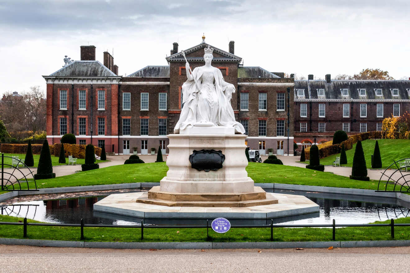 A large white statue stands in front of Kensington Palace, surrounded by manicured gardens and a small reflecting pool on a cloudy day.