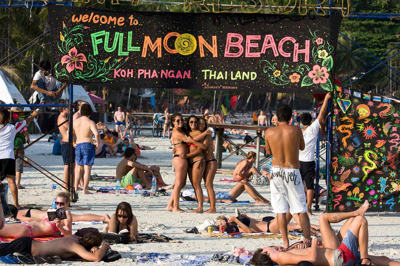 A bustling beach scene at Full Moon Party in Koh Phangan, with a welcome sign decorated in vibrant colors and designs, and tourists relaxing on the sand