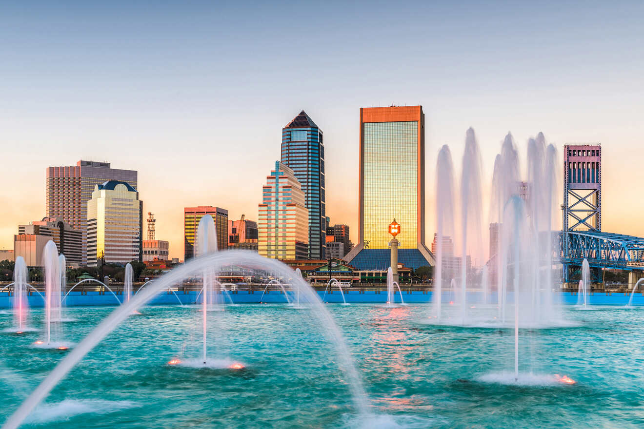 Skyline of a city with tall buildings at sunset, featuring a waterfront with multiple fountains in the foreground.