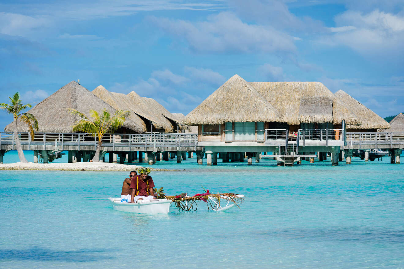 A small boat with two people and palm fronds floats on turquoise water in front of thatched-roof bungalows on stilts.