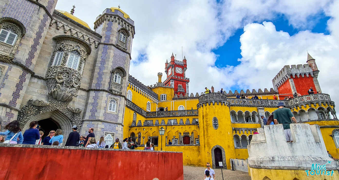 Colorful Pena Palace with red, yellow, and gray towers. Tourists explore the courtyard under a partly cloudy sky.