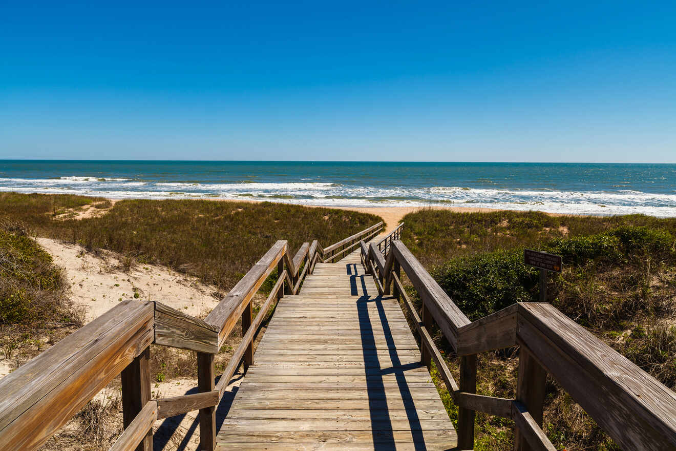 Wooden boardwalk leads to a sandy beach with waves under a clear blue sky.