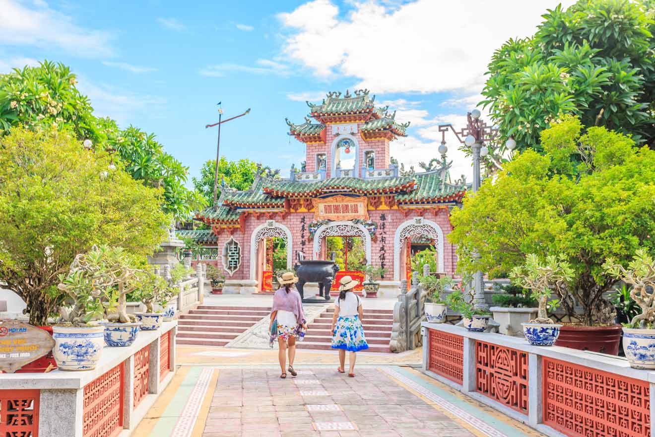 Two people walk towards an ornate, traditional Asian temple gate surrounded by lush greenery and vibrant potted plants under a bright, partly cloudy sky.