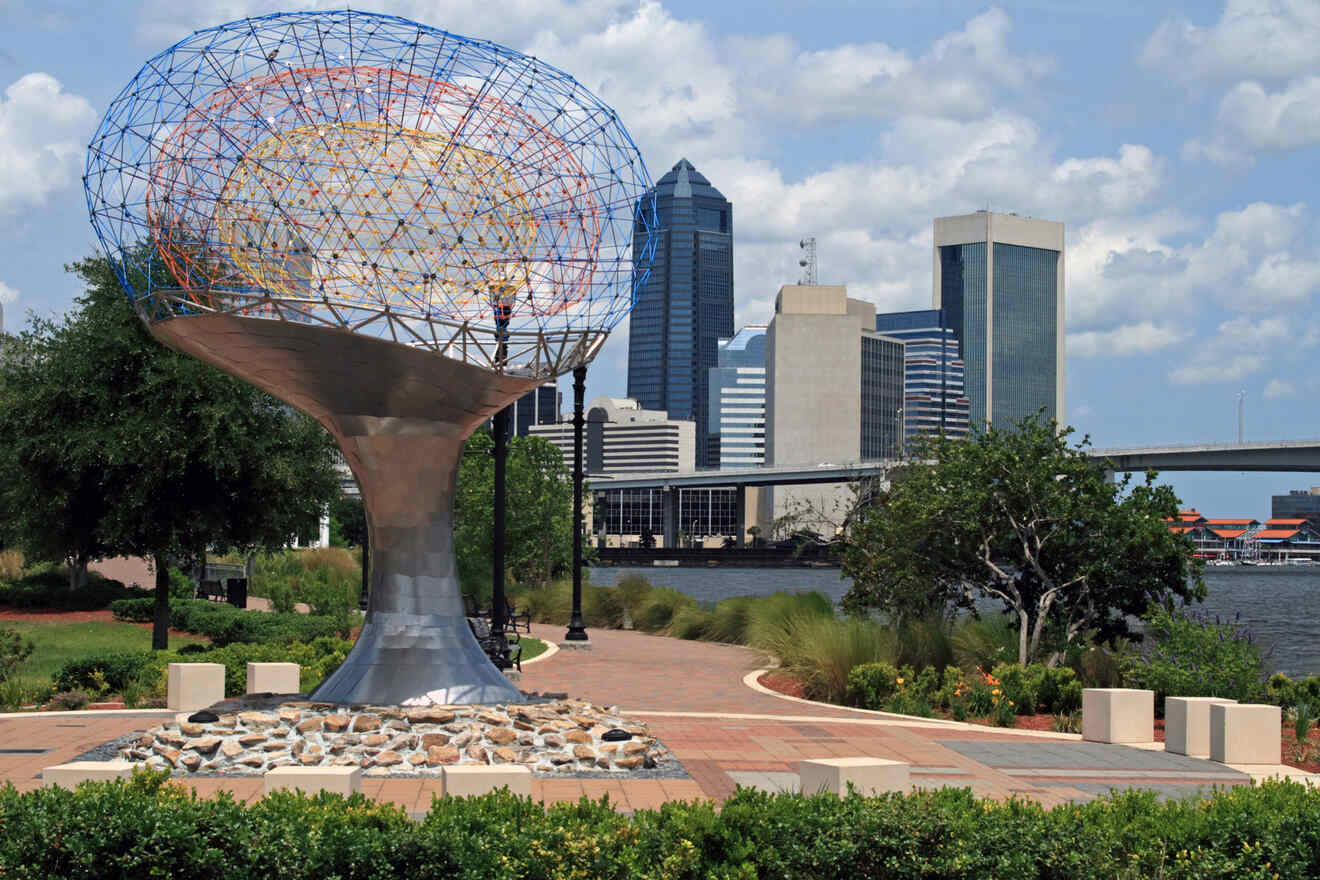 A metal sculpture with a colorful wireframe top stands in a riverside park, with city skyscrapers in the background.