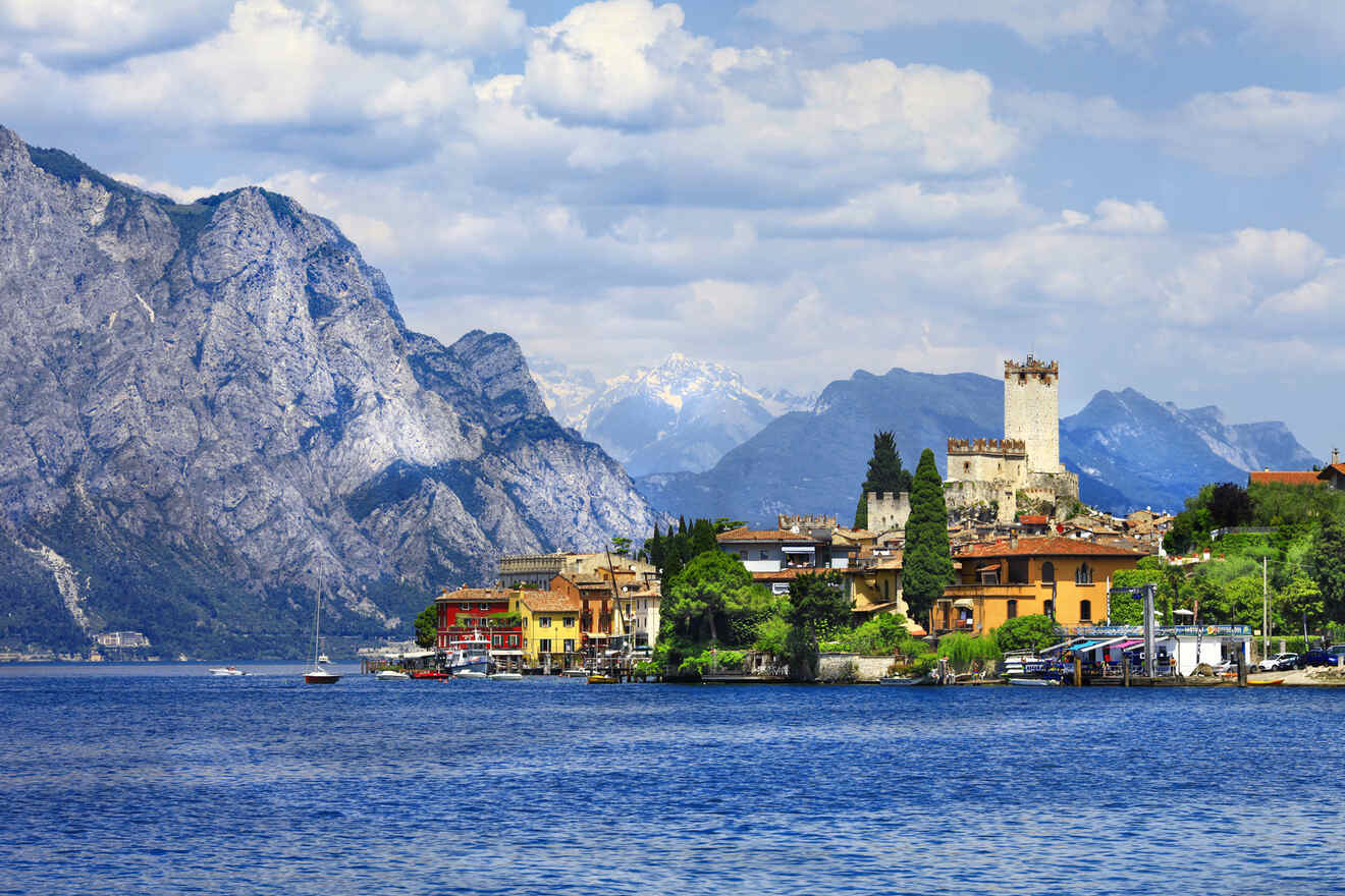 A picturesque view of Lake Garda with a historic castle and colorful buildings along the shoreline, surrounded by mountains under a partly cloudy sky.