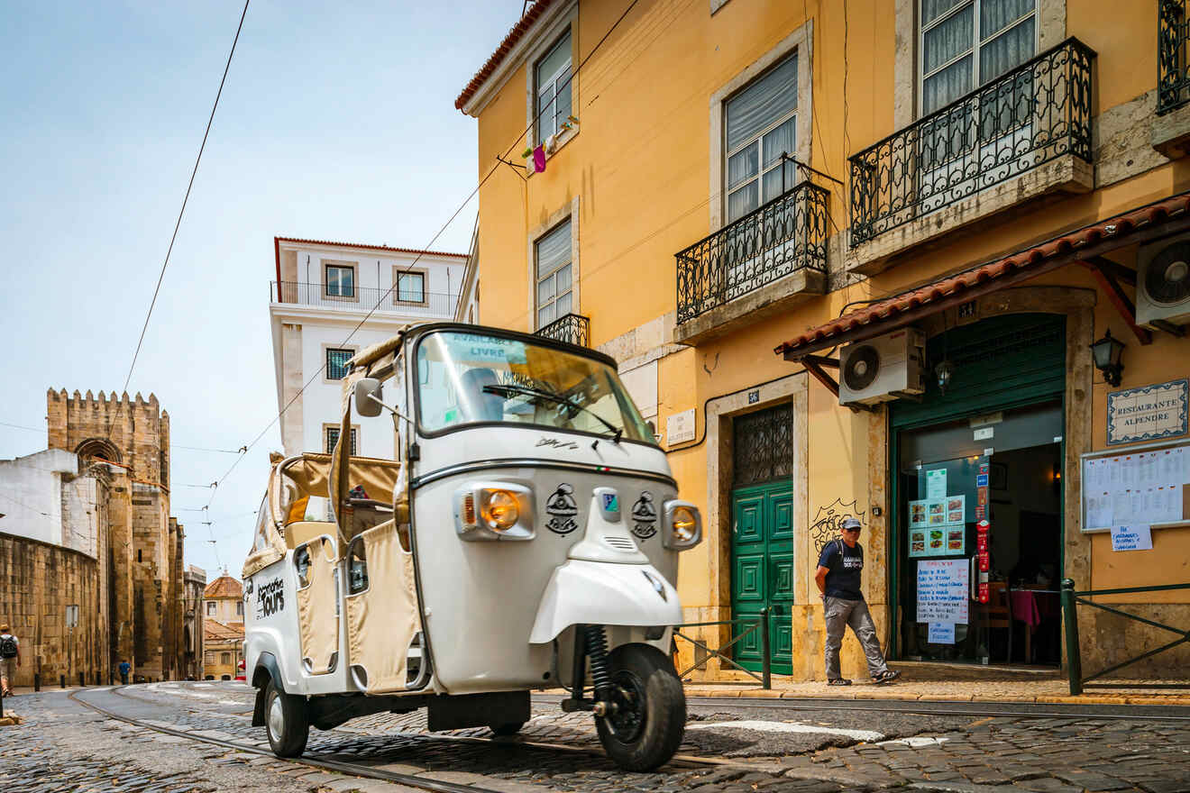 A white tuk-tuk drives on a cobblestone street in a European city, passing by historical buildings and a church in the background.