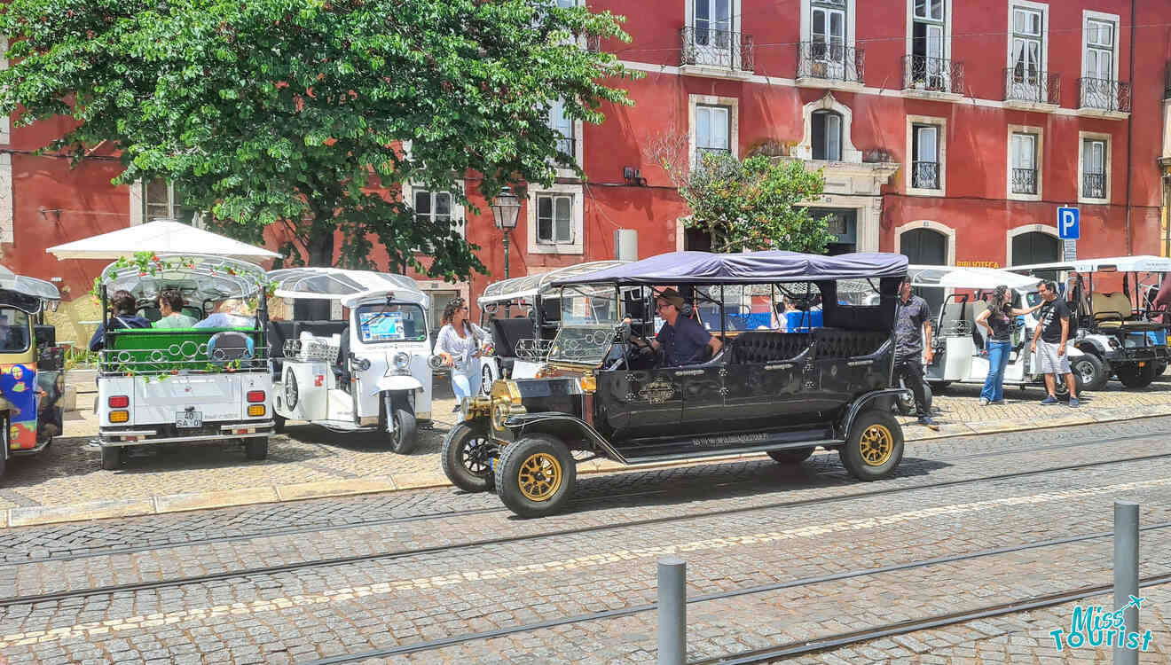 A vintage black car drives along a cobblestone street lined with parked tuk-tuks and pedestrians. A red building with white-trimmed windows and a tree are in the background.