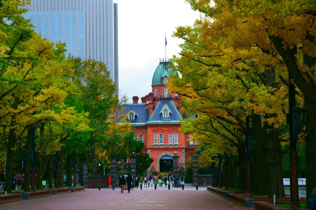 A historic red brick building framed by trees with autumn foliage, set against a backdrop of a modern skyscraper.