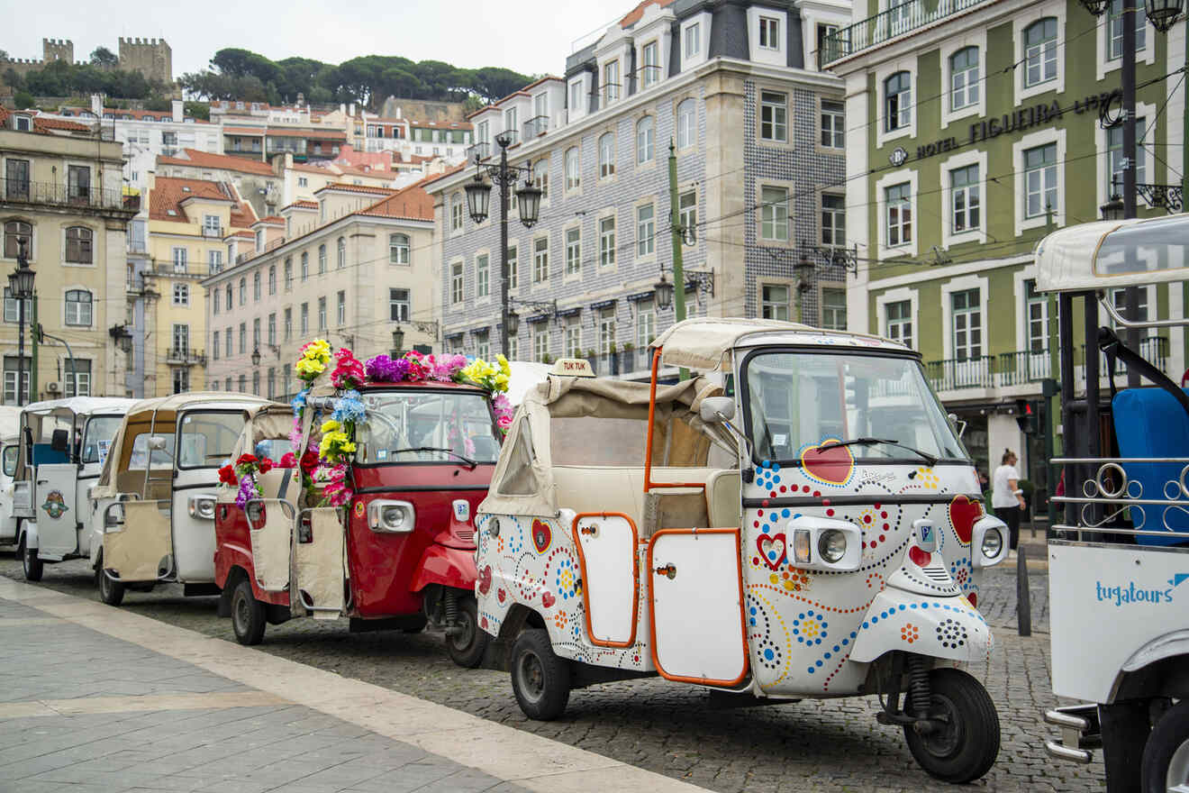 A row of decorated tuk-tuks is parked along a cobblestone street in a European city, with colorful flowers adorning one of the vehicles. Historic buildings are in the background.