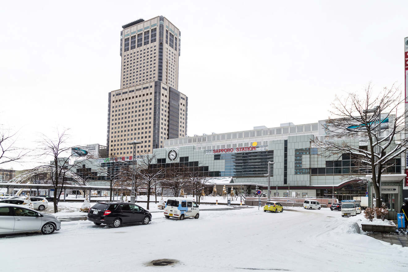 A bustling city center with a high-rise building and Sapporo Station, surrounded by snow-covered streets and parked cars.