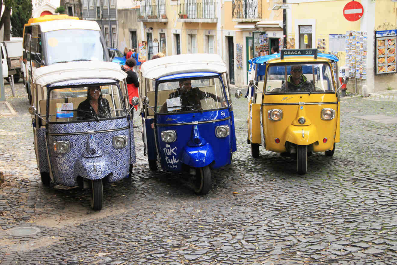 Three motorized tuk-tuks, one patterned, one blue, and one yellow, are parked side by side on a cobblestone street in an urban area. Two drivers are visible behind the windshields.