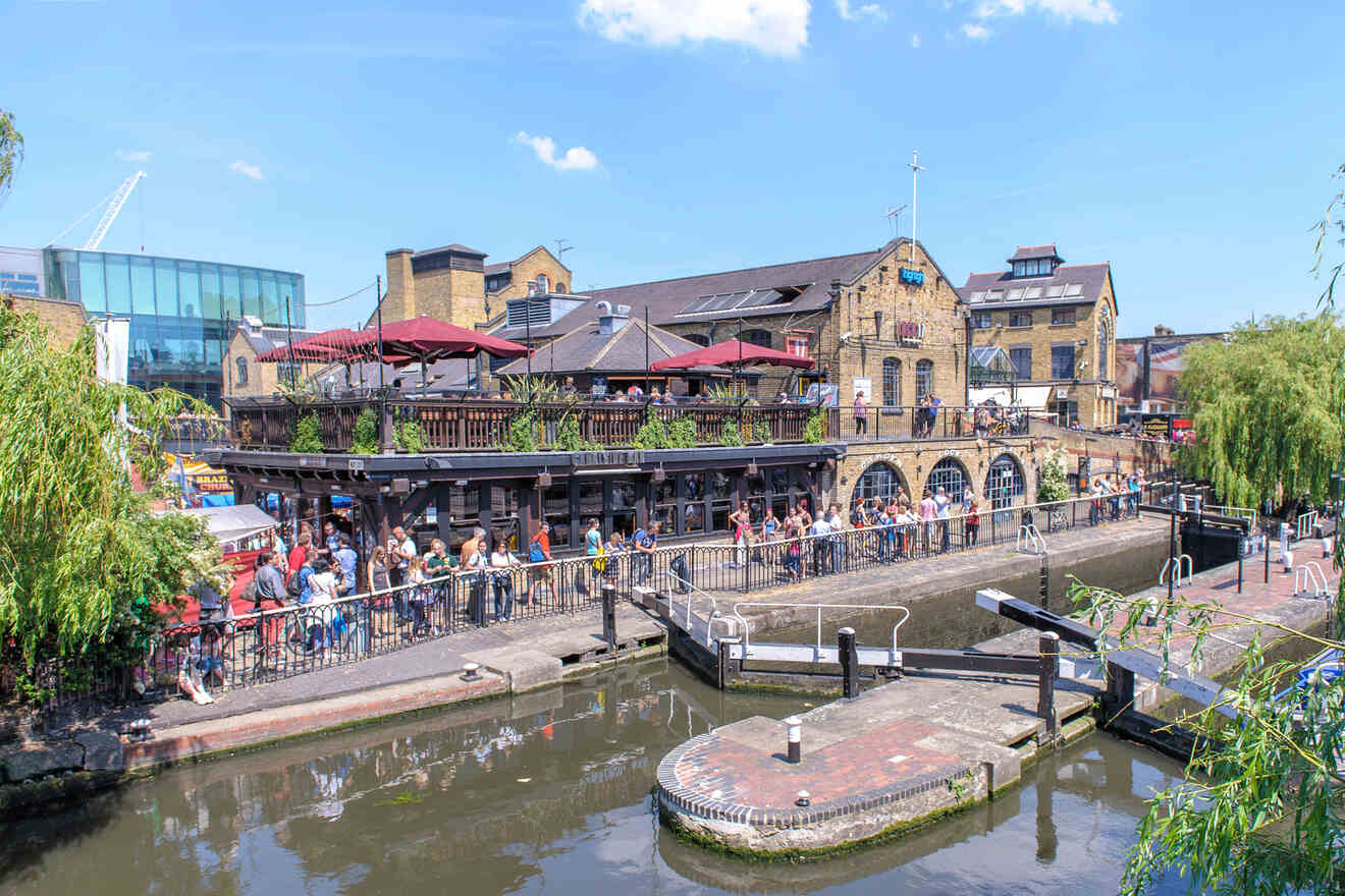 People gather around the vibrant outdoor area of Camden Market by the canal on a sunny day, with brick buildings and lush greenery in the background.