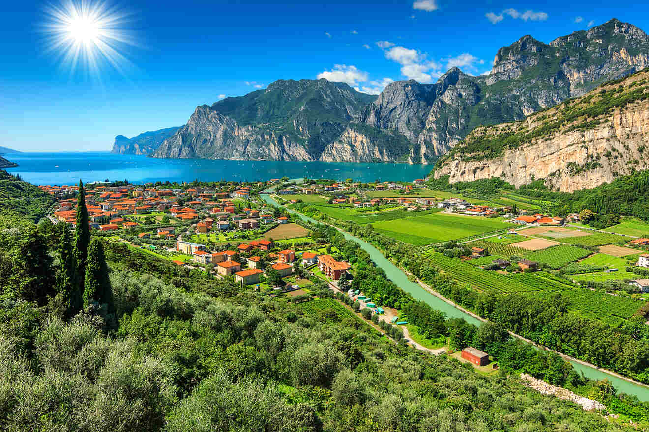 Scenic view of a vibrant village surrounded by lush greenery, a winding river, and mountains under a bright blue sky with a shining sun.