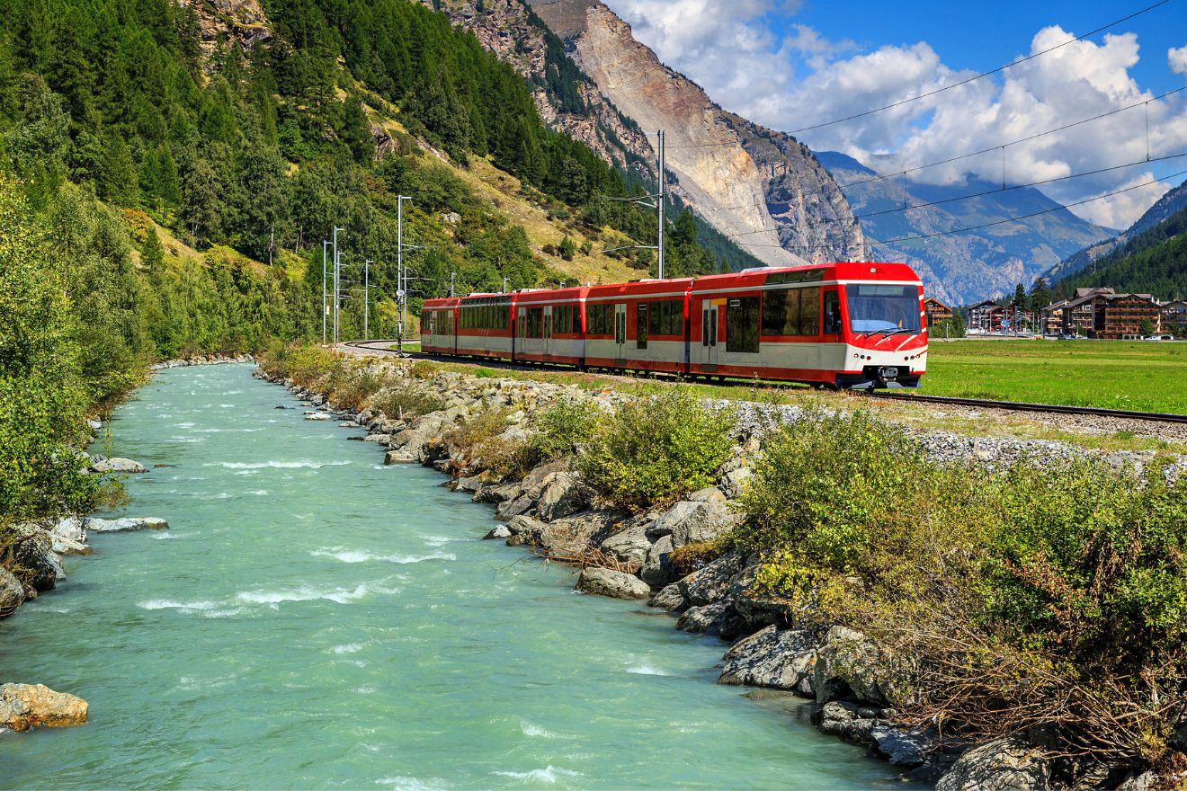 A red train travels along a track beside a green river, bordered by forested hills and mountains in the background under a partly cloudy sky.