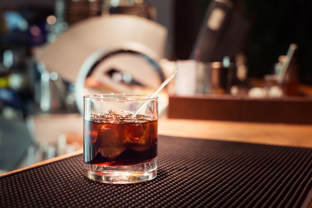 A glass containing a dark beverage with ice cubes and a stirring rod is placed on a black textured mat in a bar setting.