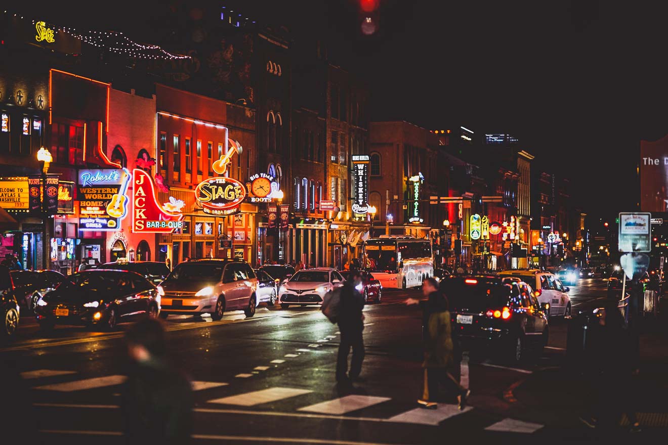 Vibrant nightlife scene on Nashville's Broadway with neon signs, bustling traffic, and pedestrians crossing the street